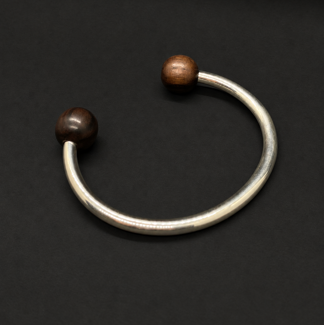 A silver-toned open cuff bracelet with two dark brown wooden beads at the ends, displayed on a solid black background.