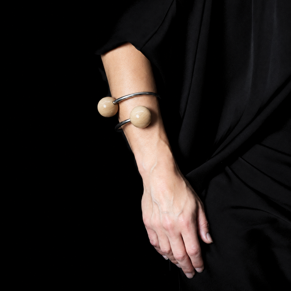 A close-up of a person's forearm and hand wearing a thick silver bangle, each end with a large smooth wood  bead, against a black background.