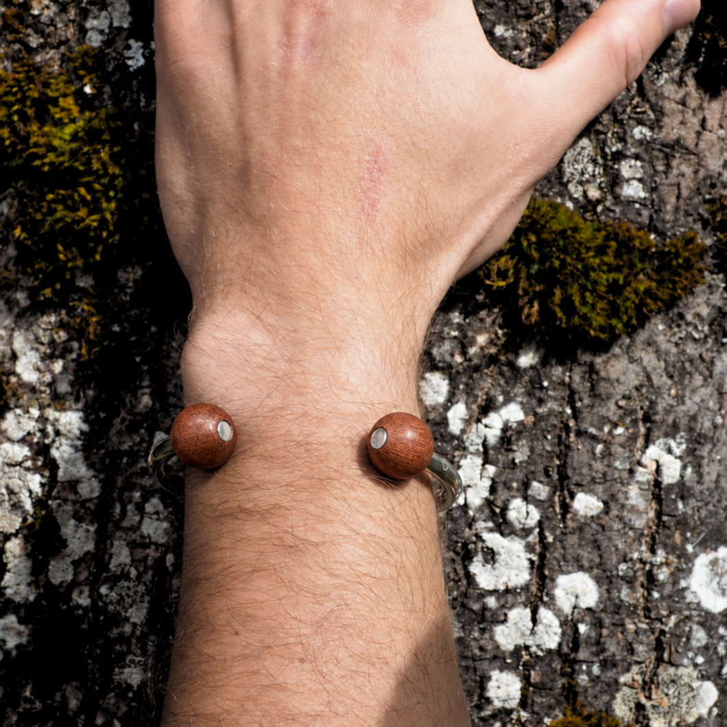 A close-up of a person's wrist and hand resting against textured tree bark, wearing two small round brown bead-like jewelry pieces on the wrist.
