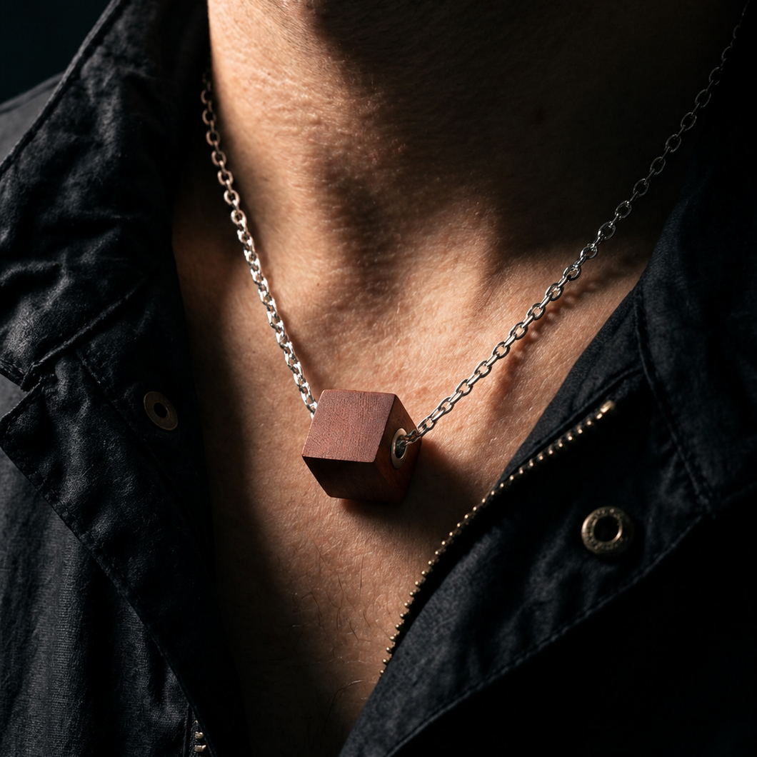 A close-up of a person's neck and chest wearing a silver chain necklace with a small cube-shaped wooden pendant, paired with a black zip-up jacket.