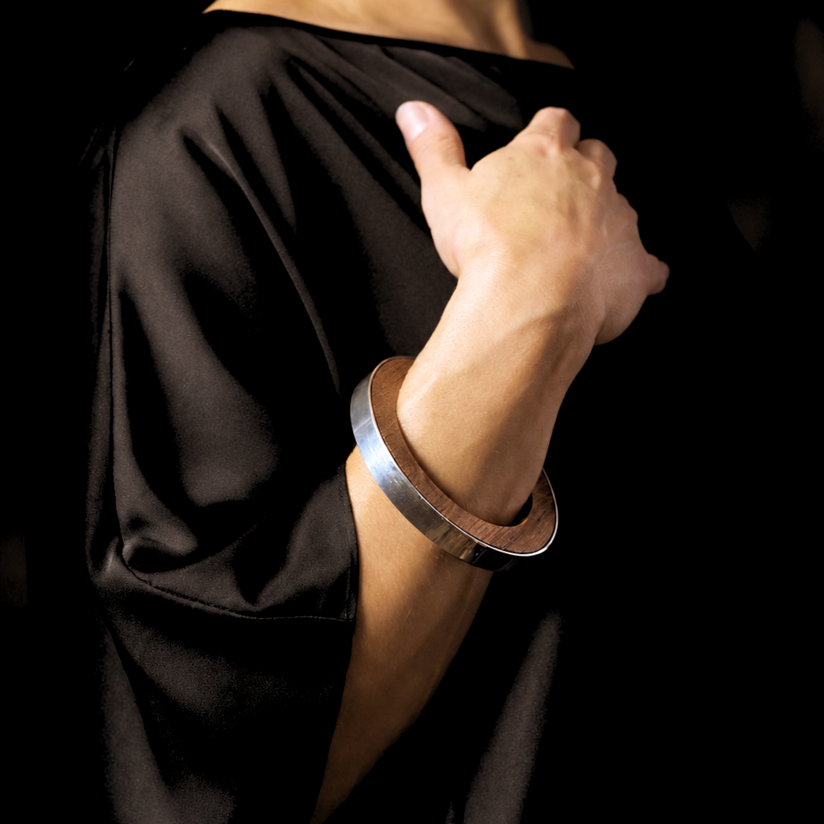 A close-up of a person wearing a wide, polished silver and wood bracelet on their wrist, dressed in a black satin or silk top against a dark background.