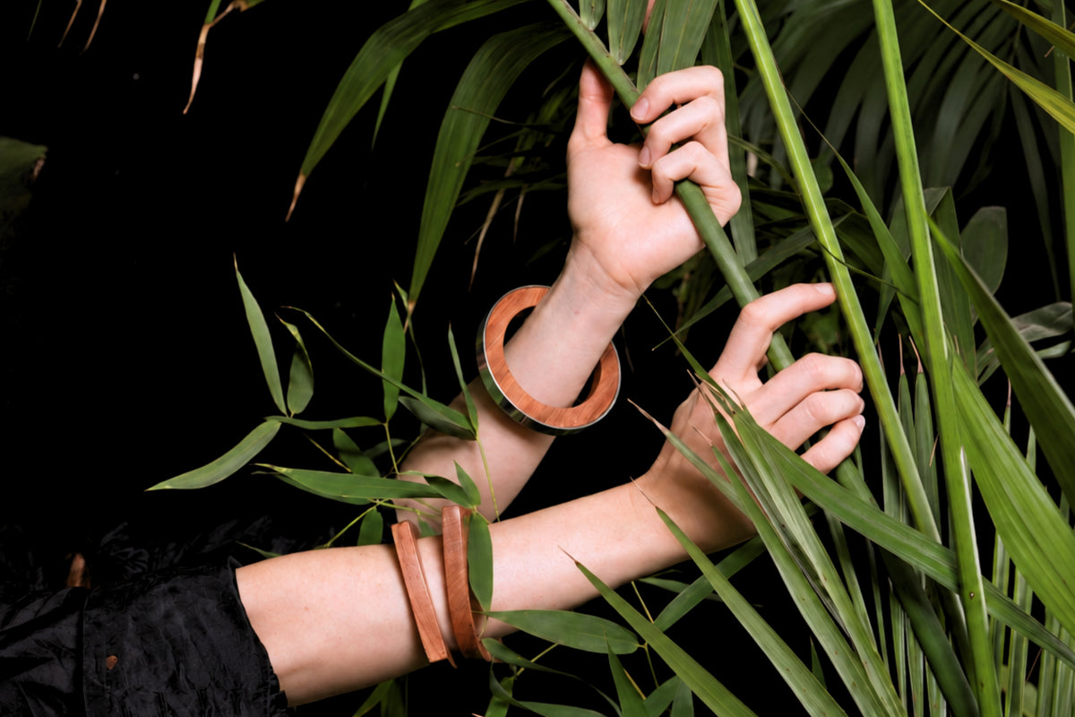 A person's hands and forearms are shown wearing two wide, circular tan leather bracelets while reaching toward green palm leaves against a dark black background.