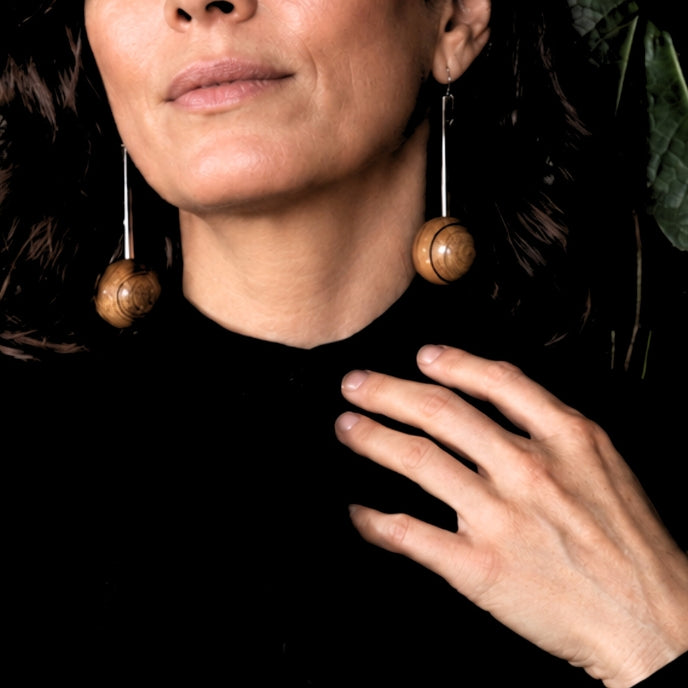 A close-up of a woman wearing long, dangling earrings with round wooden beads suspended from thin silver-toned wires, against a dark background.
