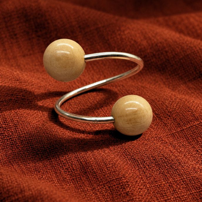 A silver  bangle bracelet with two round, light beige wooden beads, displayed on a textured rust-red fabric background.