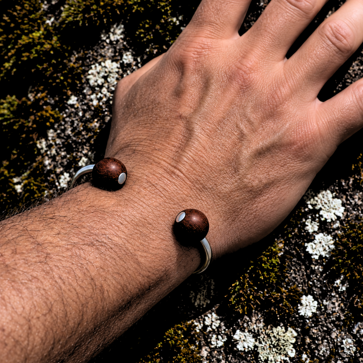 A close-up of a person's hand resting on mossy rock, wearing a minimalist silver-toned bracelet with two dark brown wooden beads.