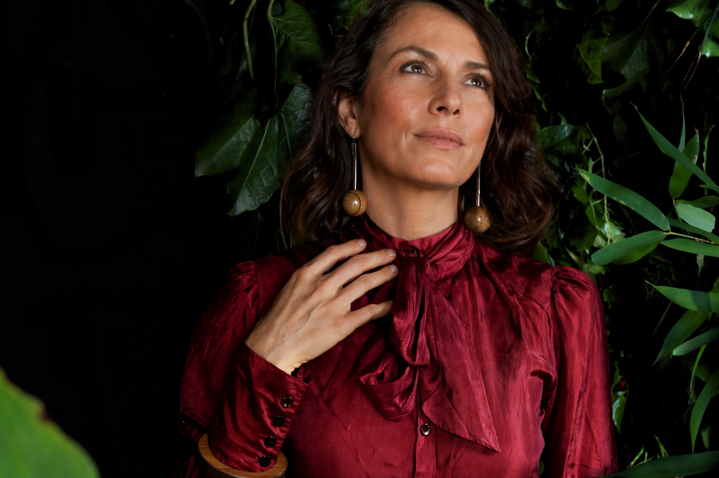 Woman wearing a red blouse and beautiful wood and silver earrings against a dark background with green leaves.