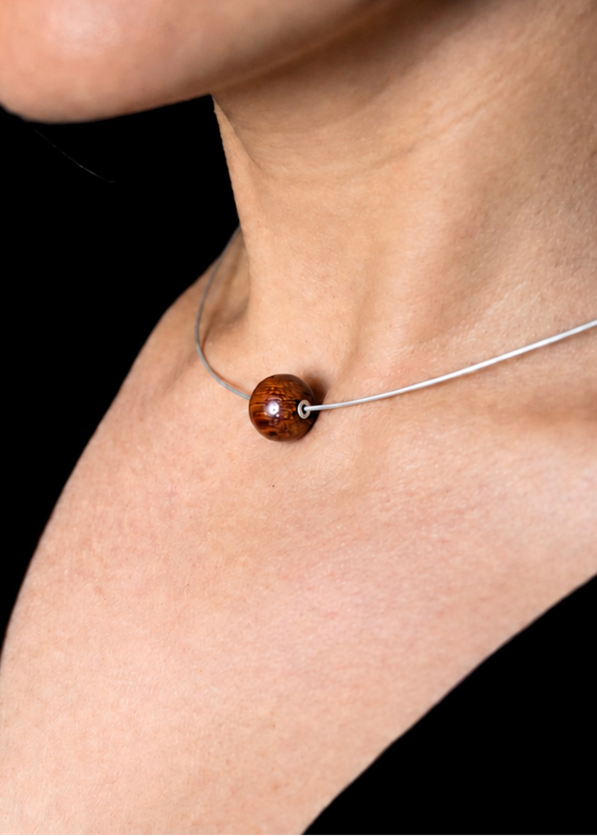A close-up of a person's neck and collarbone wearing a delicate silver wire necklace with a single polished brown wooden bead pendant.