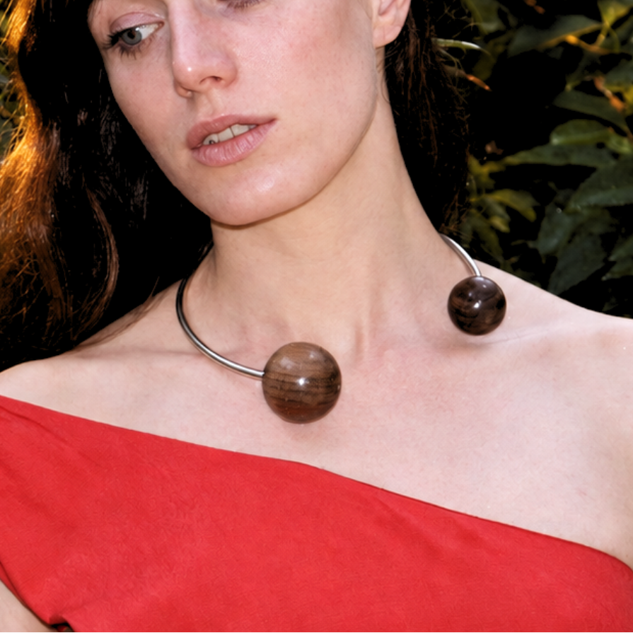 A woman with dark hair wearing a red off-the-shoulder top and a silver necklace with two large brown spherical beads, posing outdoors against a background of green leaves.