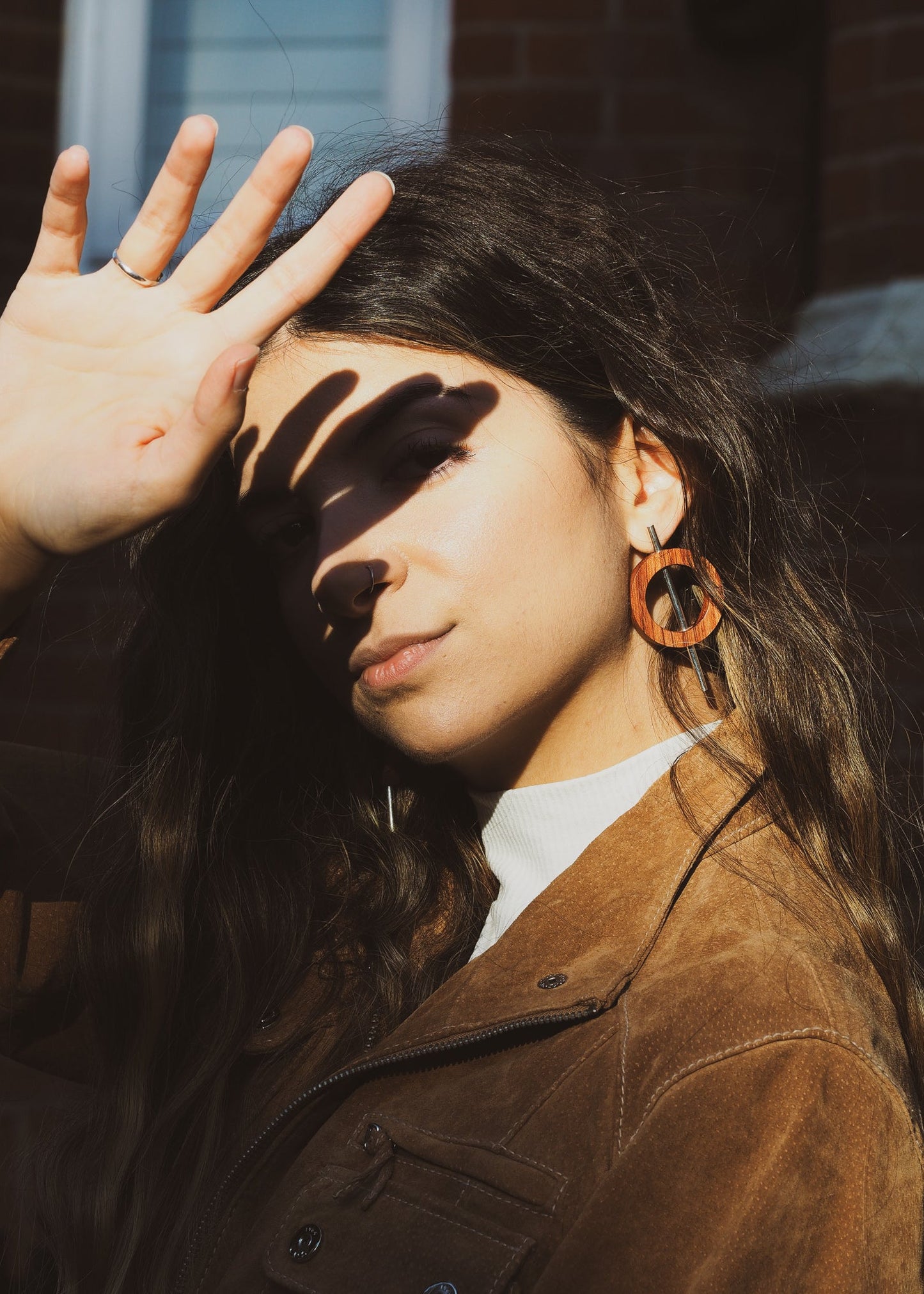 Woman with long hair and brown jacket holding hand up to face, wearing large wood and silver earrings.