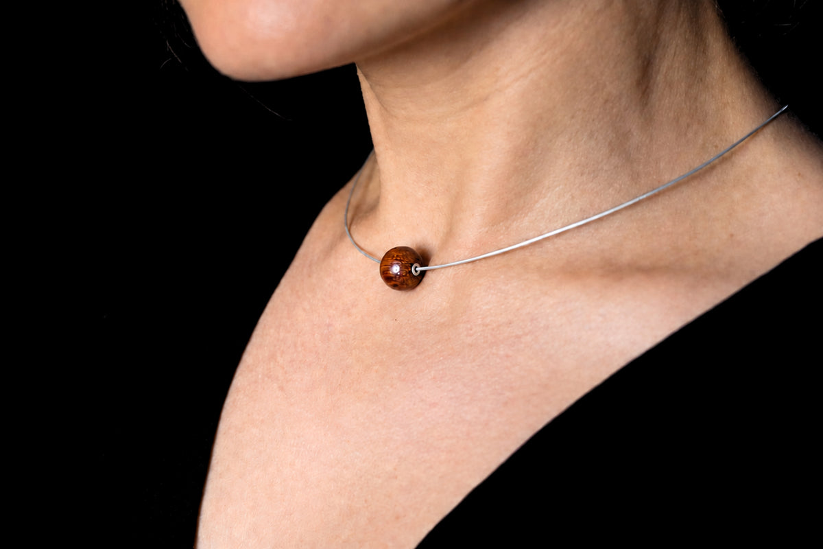 A close-up of a person's neck and upper chest wearing a delicate silver necklace with a single round brown bead pendant, set against a solid black background.