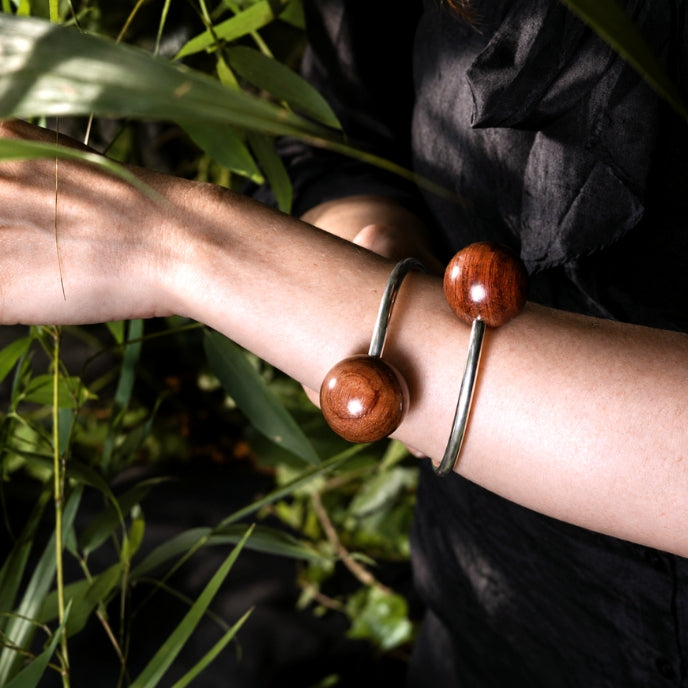 A close-up of a person's forearm wearing silver cuff bracelet,  featuring a polished reddish-brown wooden sphere, set against a background of green bamboo leaves.