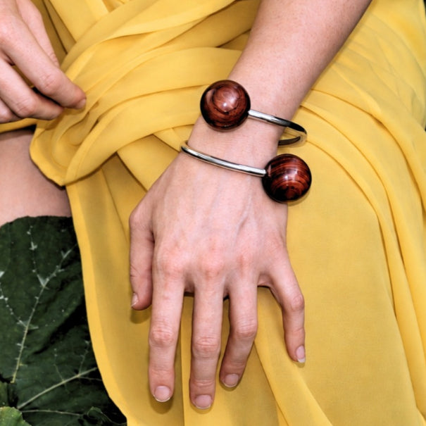 A close-up of a person's wrist wearing two silver cuff bracelets, each featuring a round polished reddish-brown wooden bead, set against a flowing yellow fabric background.