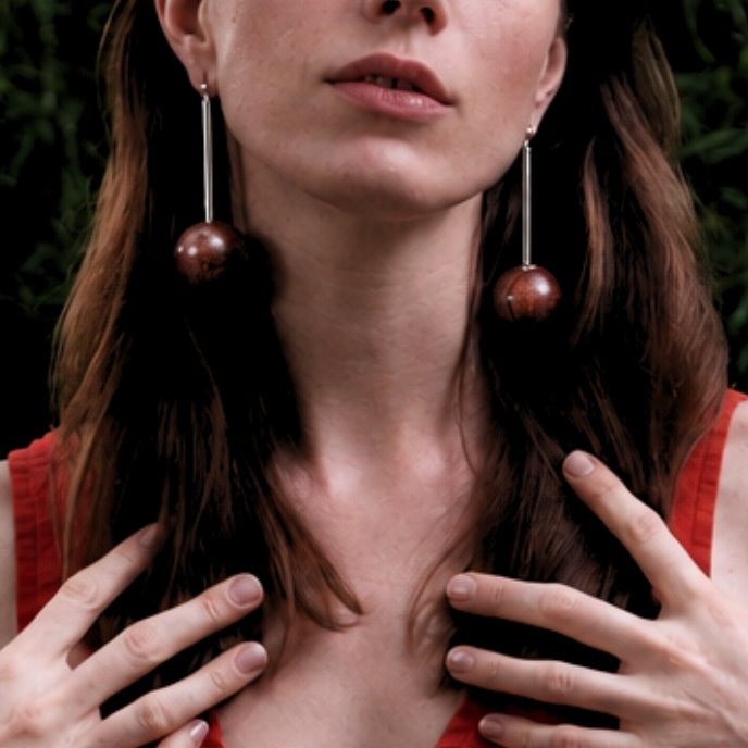 A close-up lifestyle shot of a woman wearing long dangle earrings with dark brown spherical beads and silver-toned metal wires, paired with a red top against a dark background.